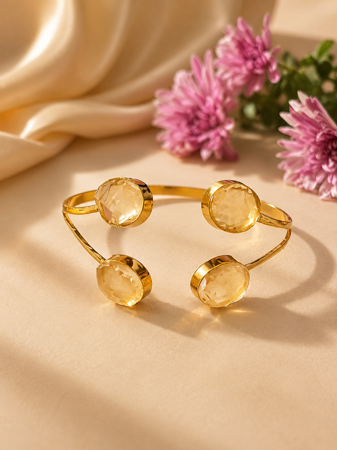 Gold open cuff bracelet with four faceted round yellow gemstones, styled on a beige satin surface with pink flowers in the background. Barkat four stone oval citrine open cuff bracelet in 18K gold plated brass by House of AAKAASHI