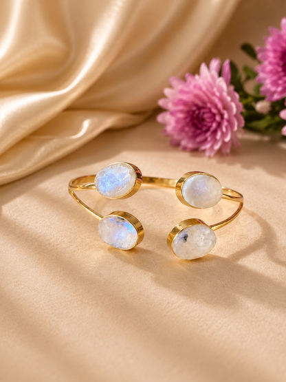 A gold-toned open cuff bracelet with four oval iridescent moonstone cabochons, styled on a beige satin fabric with pink flowers in the background. Roshan four stone oval rainbow moonstone open cuff bracelet in 18K gold plated brass by House of AAKAASHI